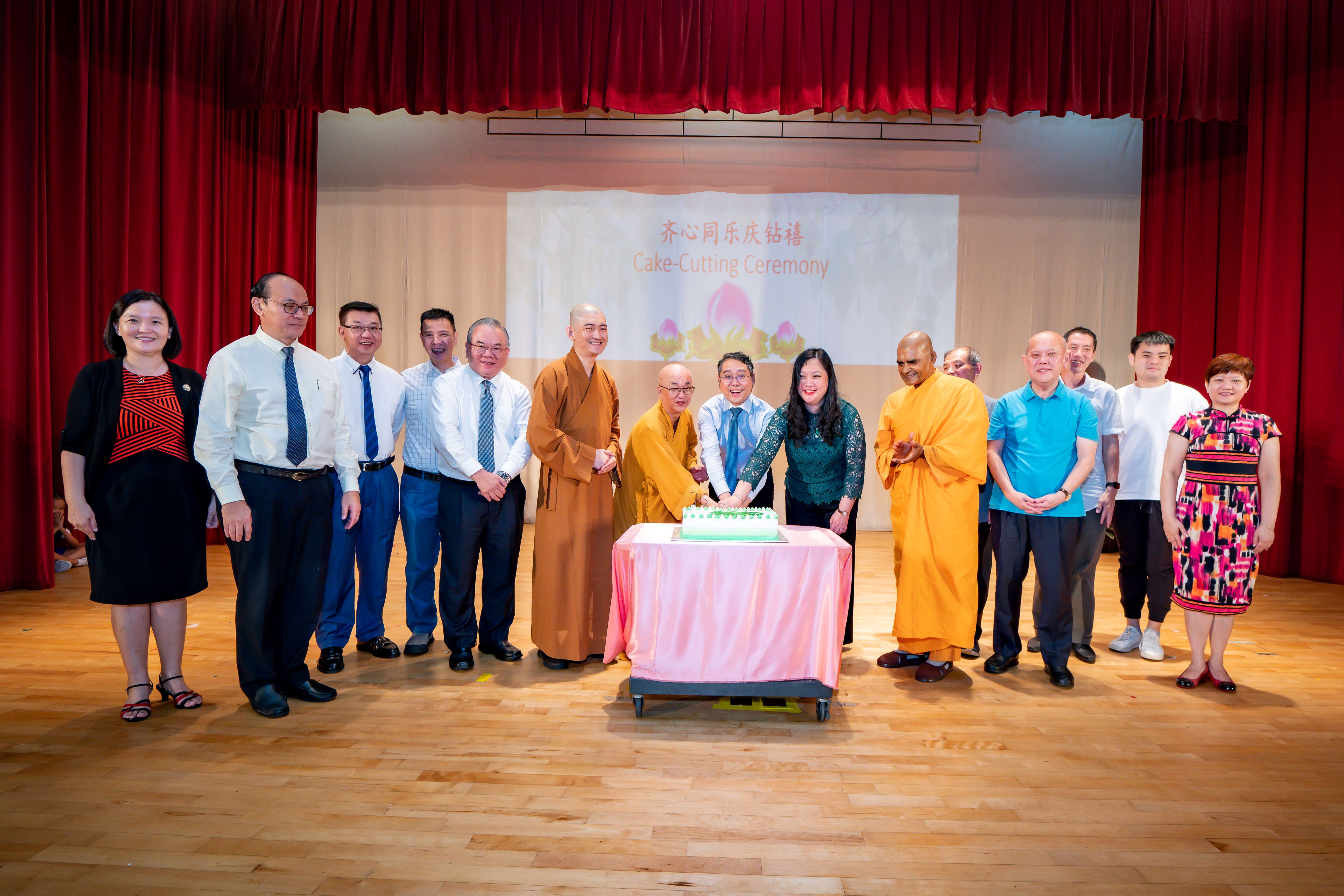 A group of people including Buddhist monks in orange robes gather on a stage for a cake-cutting ceremony.